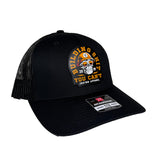 A black trucker hat with a curved brim, black mesh back, and a circular embroidered patch on the front. The patch features a skull wearing a yellow helmet with the text 'BUILDING SHIT YOU CAN'T' and 'JRNYMN APPAREL'.