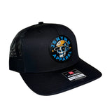 Black trucker hat with a circular embroidered patch on the front featuring a skull in a firefighter hat and the text 'JRNYMN APPAREL'. The hat has black mesh back panels and a black curved brim