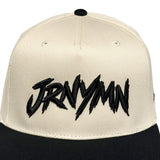 A structured baseball cap with an off-white crown and a black flat brim, featuring the word 'JRNYYMN' embroidered in a black jagged font across the front.
