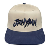 A structured snapback cap with an off-white crown and a navy blue flat brim. The front of the cap features a stylized, jagged navy blue embroidered logo that reads 'JRNYMN'.
