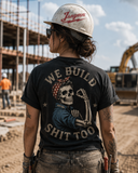 A woman on a construction site seen from behind, wearing a black graphic t-shirt with a large back print of a skeleton worker and the text 'WE BUILD SHIT TOO', a white hard hat with red script, and sunglasses.